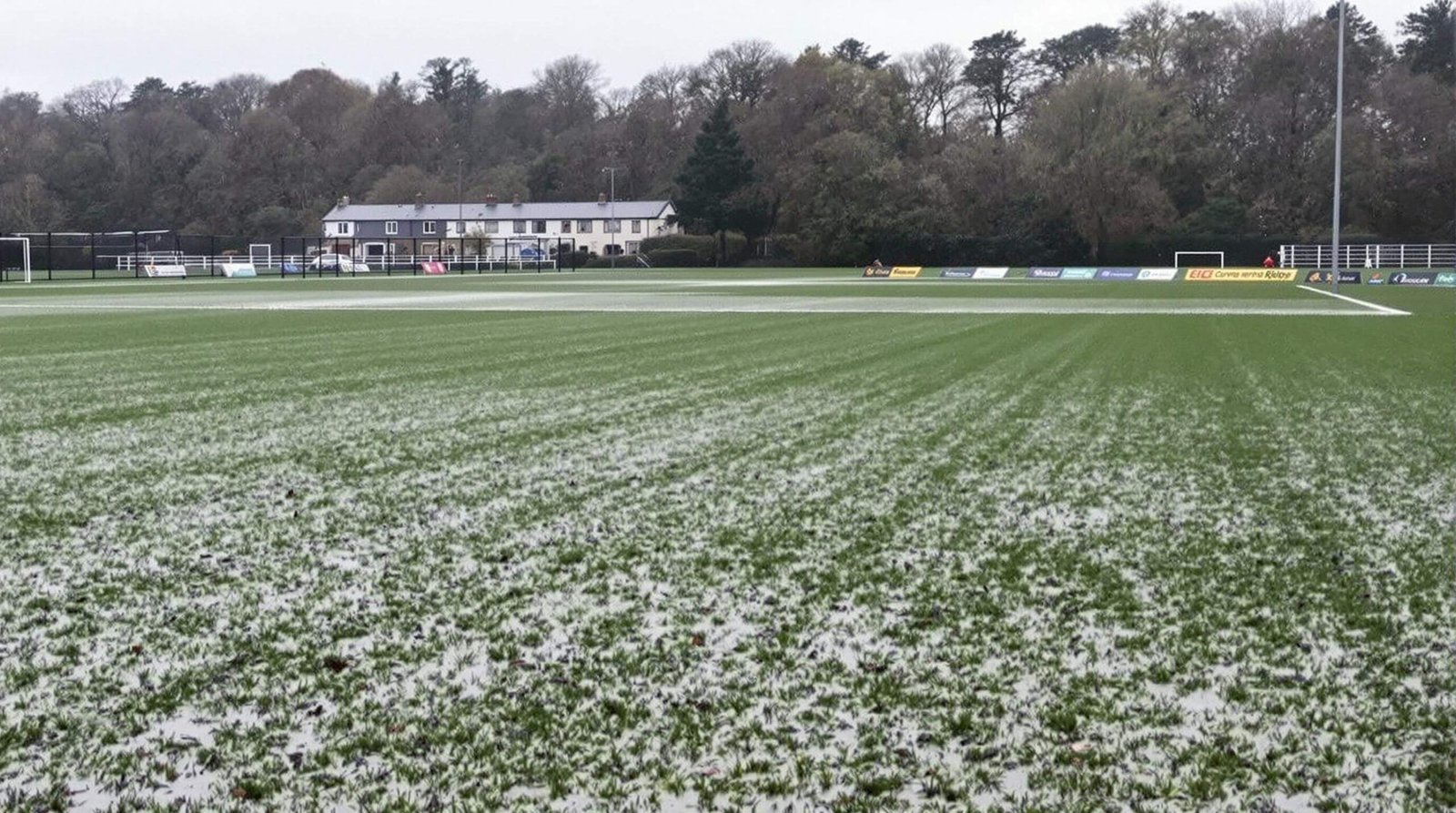 "Waterlogged rugby pitch at Footes Lane, Guernsey with standing puddles, empty grandstand, and 'Match Postponed' sign"